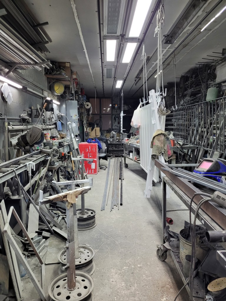 Picture of a metal worker's work place. Pictured are multiple tools, metal frames hanging from the ceiling, a welder's mask etc.