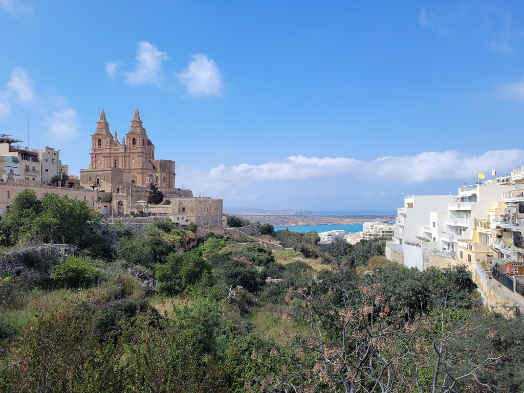 Landscape picture of a church in the city of Mellieha, Malta. Modern white buildings can be seen on the right and Mellieha Bay in the distance. 