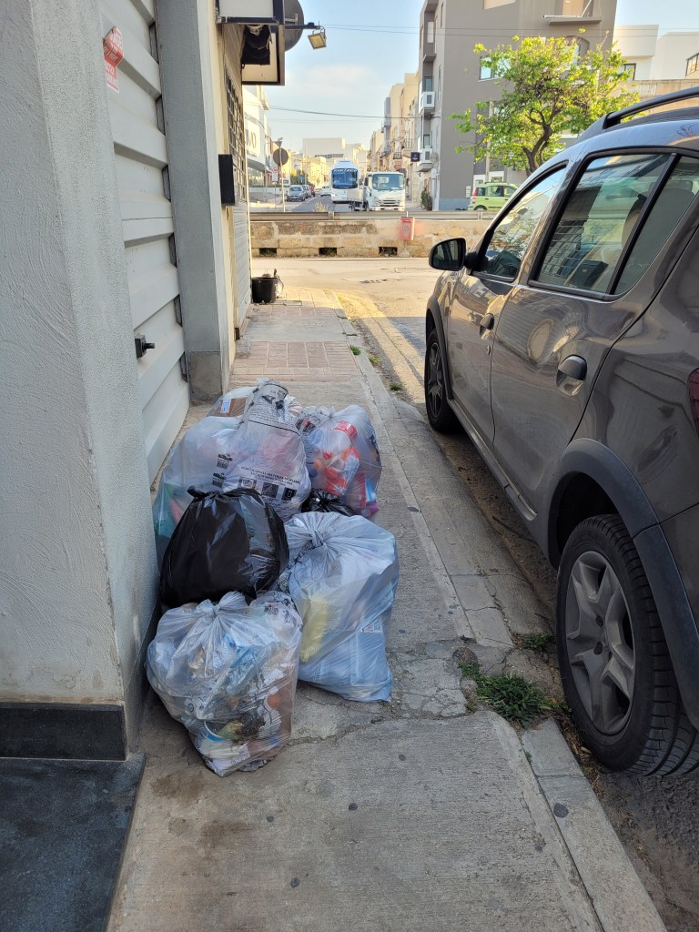Picture of a street with multiple full garbage bags laying next to a house.