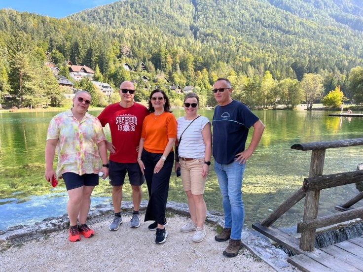 Viisi ihmistä seisoo ja poseeraa kameralle järven edessä.