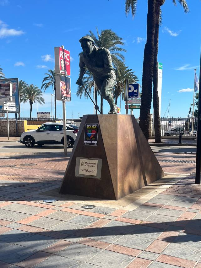 kuva Fuengirolan kalastajien kunniaksi tehdystä patsaasta, taustalla mainoskylteissä KFC:eelle, subwaylle, muille ravintoloille ja näkyvissä myös autoja tiellä.