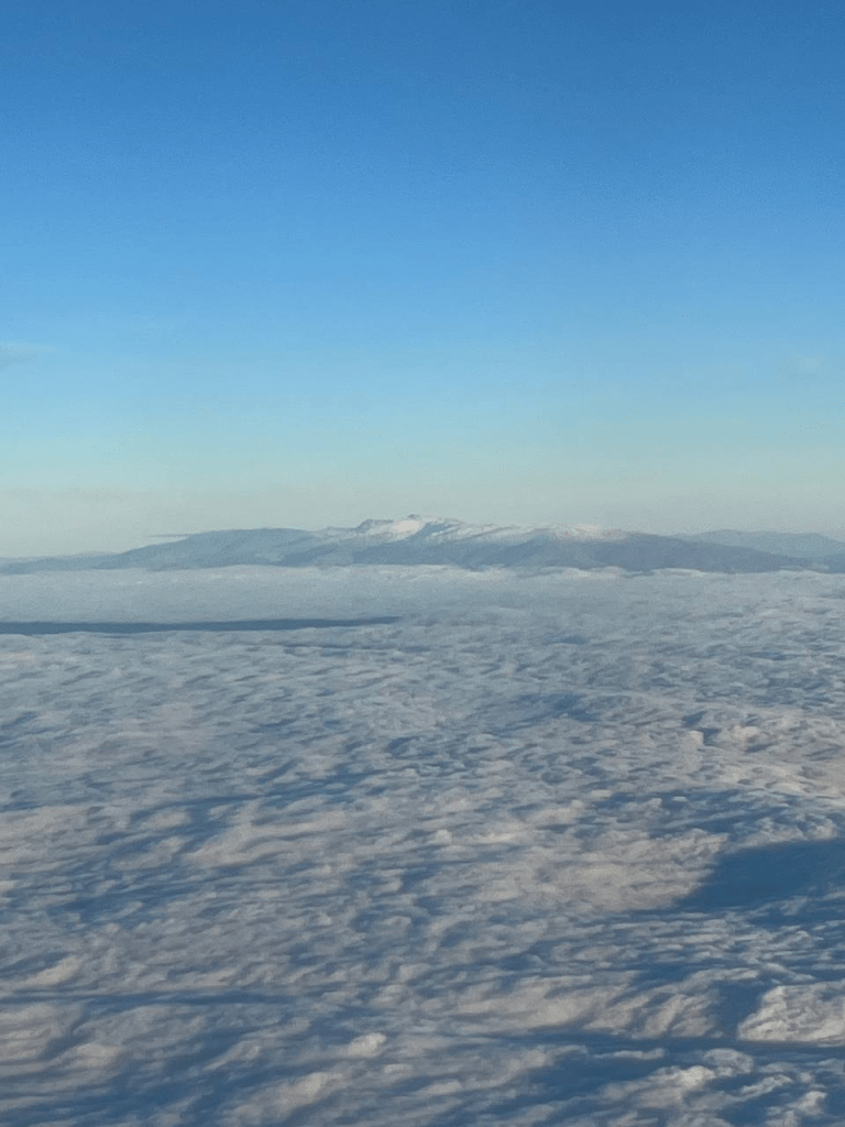 Lentokoneesta otettu kuva missä näkyy pilviä, iso vuori ja sininen taivas.