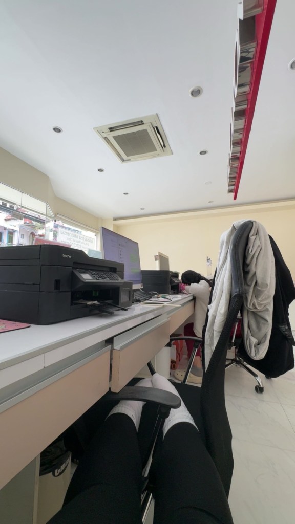 Kuva on Klinikan sisältä. Kuvassa on Klinikan vastaanottotiski jonka päällä on tulostin, tietokone ja tiskin takana istun minä ja toinen hoitaja.