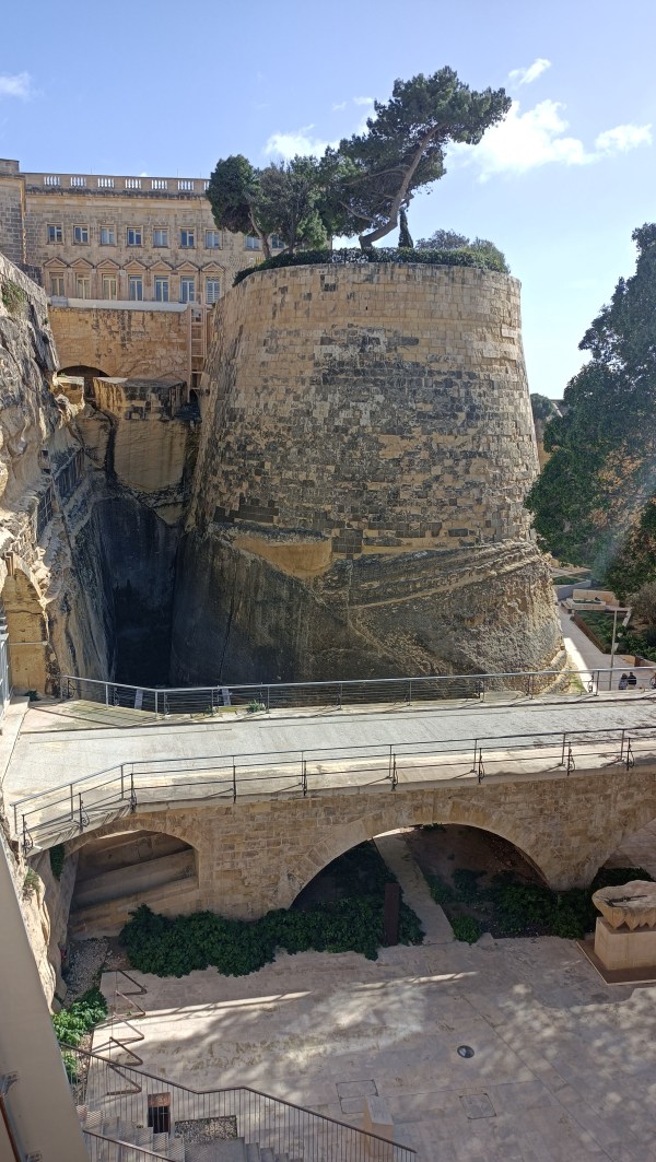 Korkea kivinen linnoitus jonka päällä puutarha ja etualalla silta