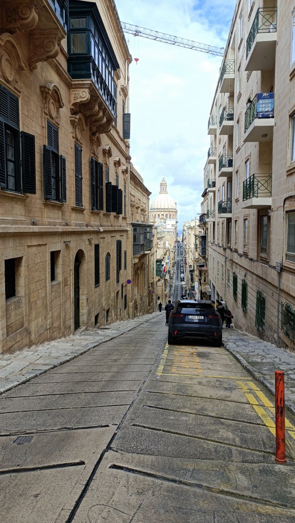 näkymä pitkästä laskevasta kapeasta kadusta jossa auto parkissa etualalla ja kirkon kupoli näkyy taustalla .