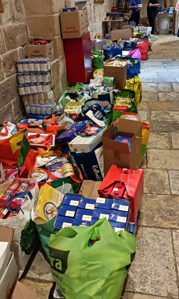 Paljon ruoka lahjoitus kasseja ja laatikoita kirkon pitkän käytävän lattialla. Taustalla kokki ja varaston vastuuhenkilö keskustelevat.